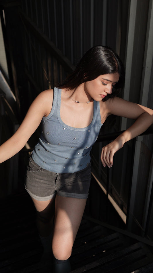 A person wearing a washed blue denim tank top with silver accents and rhinestones, paired with black shorts.