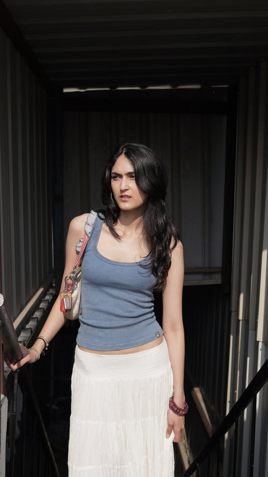 A person wearing a denim blue tank top with frills, paired with a white skirt, standing in front of a dark background.