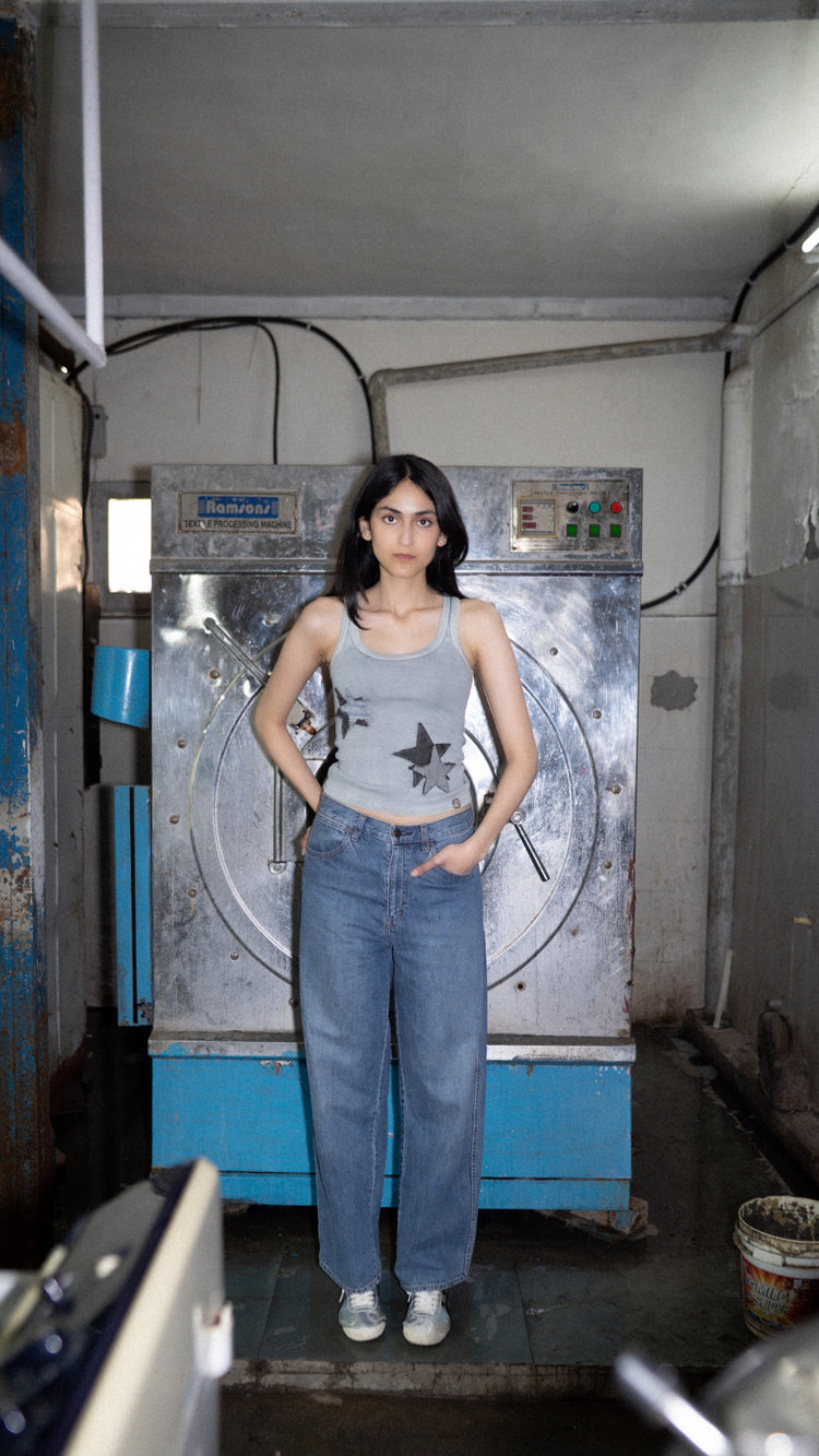 A person standing in front of industrial laundry machines wearing a gray tank top with denim and striped patches.