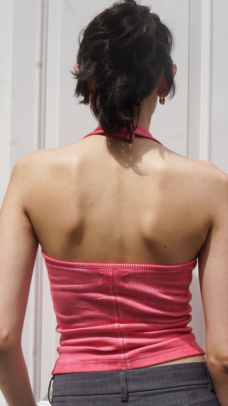 Person wearing a pink halter neck tank top and gray pants against a white wall