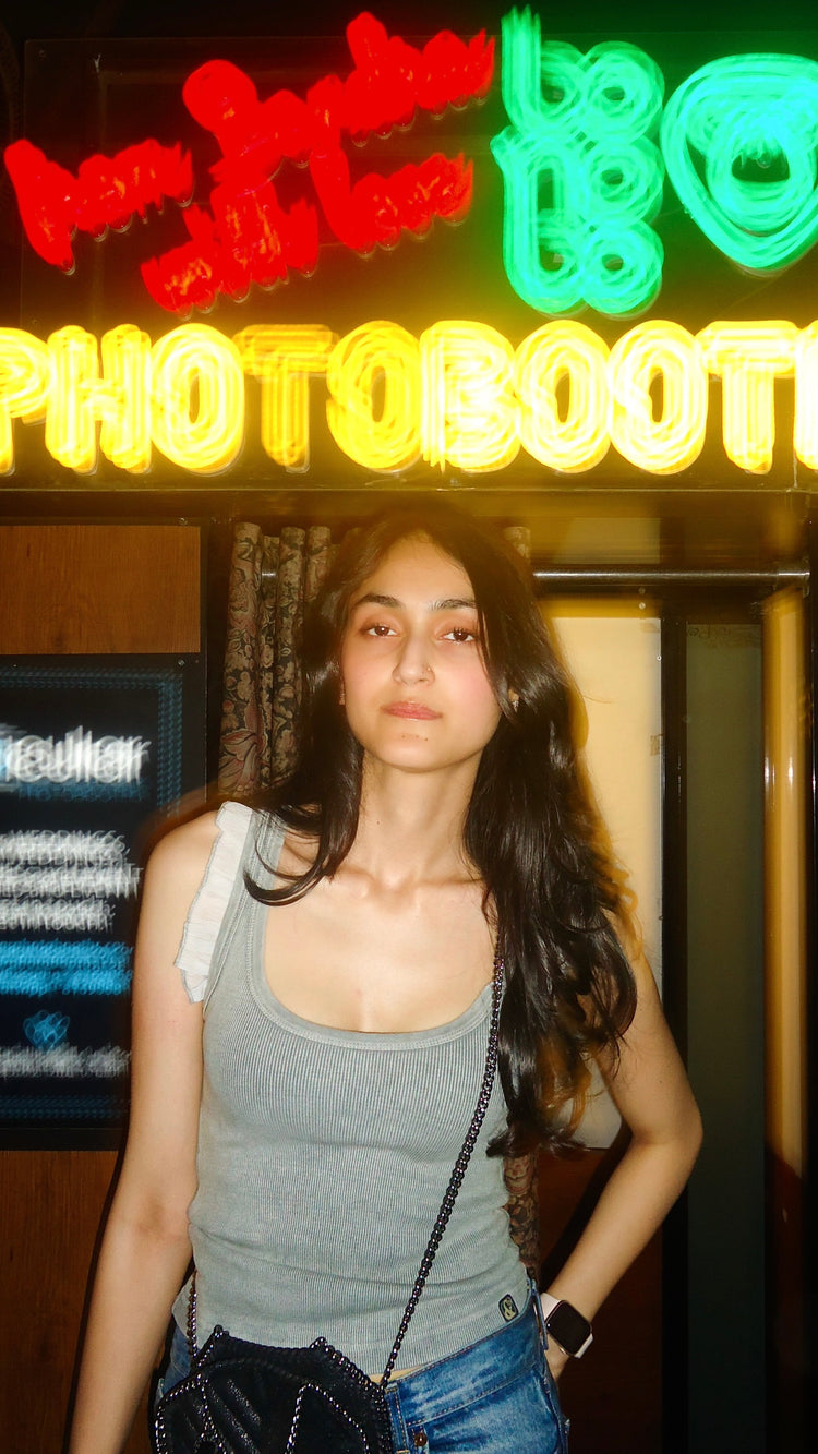 A person wearing a gray tank top with frills at the shoulder area, standing in front of a neon sign that reads "Photo Booth"