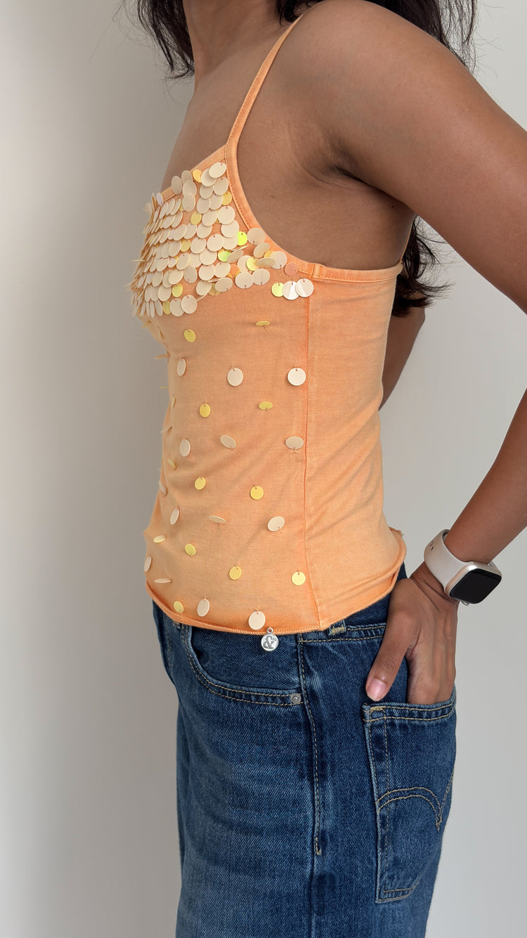 Side profile of a model wearing an orange sequin camisole top and blue jeans against a plain background