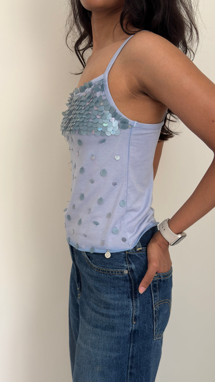 Side profile of a model wearing a light blue sequin camisole top and jeans against a plain background