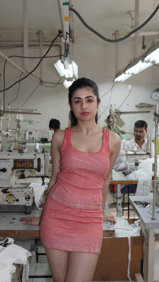 A woman wearing a shell pink sleeveless tank top with rhinestone-studded constellations and silver foil accents, with a matching skirt standing in a garment factory.